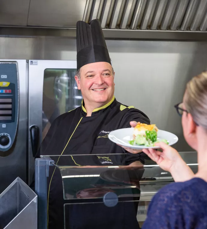 Chef cuisinier d'Ansamble souriant et servant un plat à une cliente dans une cuisine professionnelle, illustrant la qualité du service et l'engagement envers la satisfaction des clients.